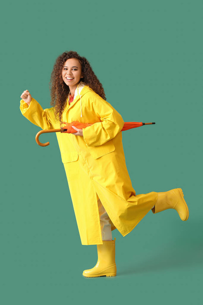 Молодая афроамериканка в жёлтом плаще и резиновых сапогах с зонтиком на зелёном фоне - Фото, изображение
