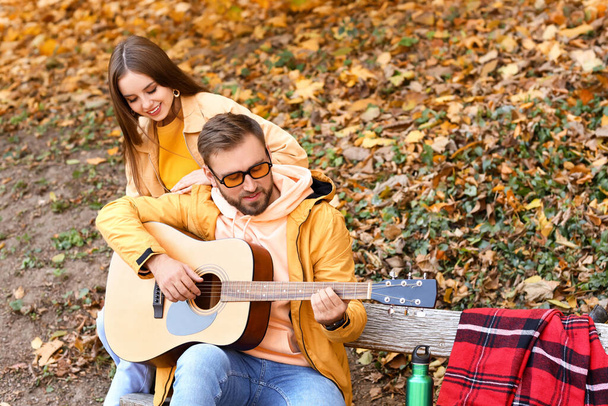 Pareja joven sentada en el banco y tocando la guitarra en el parque de otoño - Foto, imagen