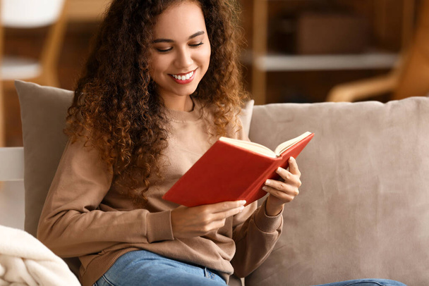 Hymyilevä nuori afroamerikkalainen nainen lukee kirjaa sohvalla kotona - Valokuva, kuva