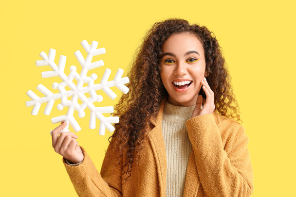 Surprised young African-American woman with big snowflake on yellow background - Foto, Imagen