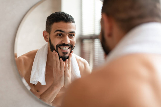 Впевнений молодий арабський хлопець, який дивиться у дзеркало, торкаючись бороди. - Фото, зображення