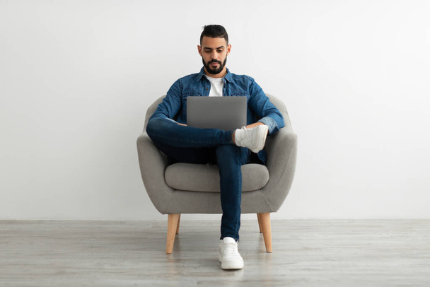 Millennial Arab człowiek siedzi w fotelu, za pomocą laptopa do pracy online lub komunikacji przeciwko białej ścianie studio - Zdjęcie, obraz