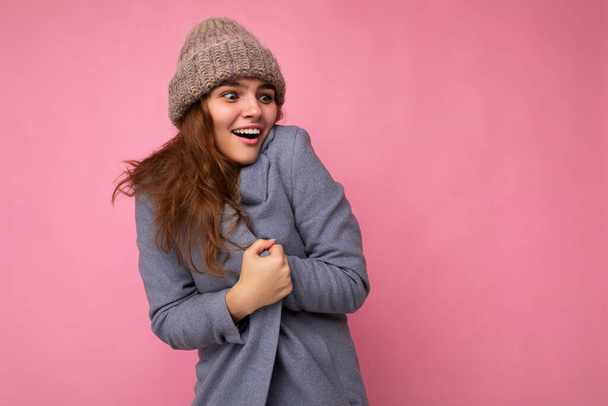 美しい幸せな若いブルネットの女性の写真ピンクの背景の壁の上に隔離グレーの秋のコートとグレーの暖かい帽子を身に着けている側を見て寒さを感じ - 写真・画像