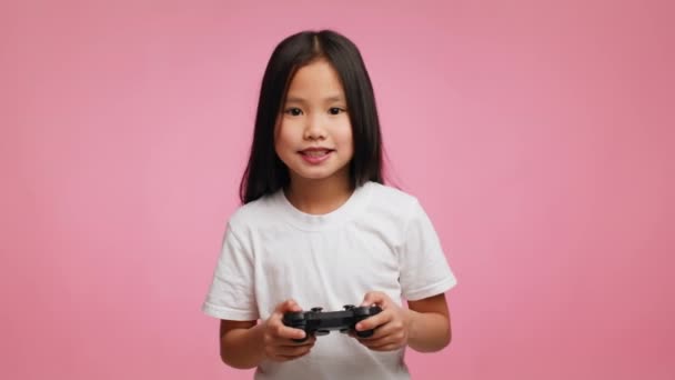 Emocionada niña japonesa jugando videojuego de pie sobre fondo rosa - Imágenes, Vídeo