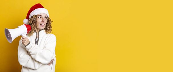 Young woman in santa claus hat with a megaphone on a yellow background. Banner - Photo, image