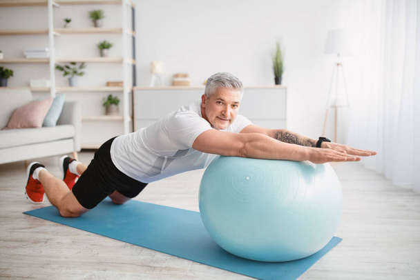 Home workout concept. Athletic senior man exercising with fitness ball, working out core muscles at home - Photo, Image