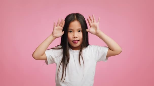 Азиатская девочка Гримасинг держа руки возле храмов, розовый фон - Кадры, видео