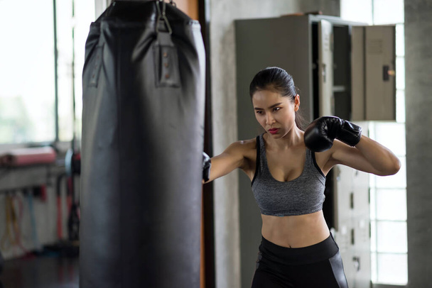 Aziatische gespierde vrouwelijke bokser in blackgloves bokstraining door het ponsen van zandzak in de fitnessruimte om Muay Thai oefenen. Professionele sport. bodybuilding en gezonde levensstijl concept. - Foto, afbeelding