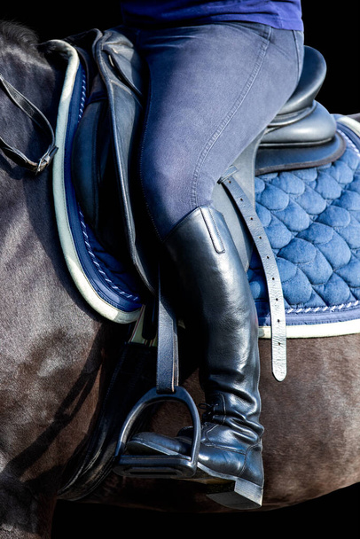A rider's foot on black horse closeup isolated on black background. A woman's booted foot standing in a black stirrup of horse saddle. - Foto, Bild