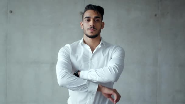 Jeune homme d'affaires arabe confiant en chemise formelle souriant à la caméra, posant avec les mains croisées, mur gris, ralenti - Séquence, vidéo