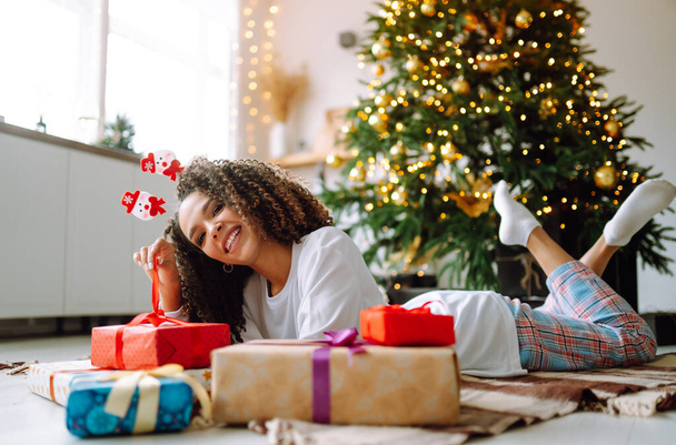 Portret van een vrolijke vrouw die de wintervakantie viert met grote geschenkdozen in de buurt van een kerstboom. Kerstverrassing. - Foto, afbeelding
