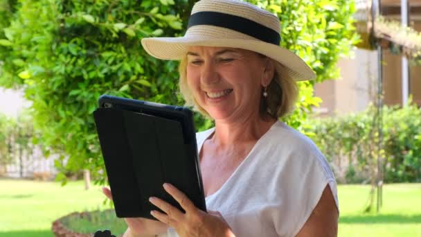 usmívající se starší blondýnka v klobouku používá tablet PC, digitální tablet pro pracovní práci nebo studium ve své vlastní zelené zahradě. žena ve věku 50-55 let. Koncept jejich vlastního podnikání a školení - Záběry, video