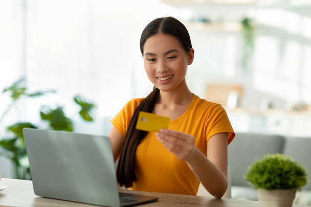 Щаслива азіатка з ноутбуком і кредитною карткою сидить за столом, купуючи товари в веб-магазині і посміхаючись, вільне місце
 - Фото, зображення