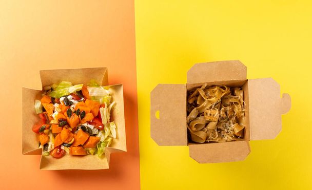 gezond voedsel. heerlijke pompoensalade met groenten in een kartonnen doos op een oranje achtergrond en pasta met kip in een kartonnen doos op een gele achtergrond. Afhaaleten. Uitzicht van bovenaf. - Foto, afbeelding