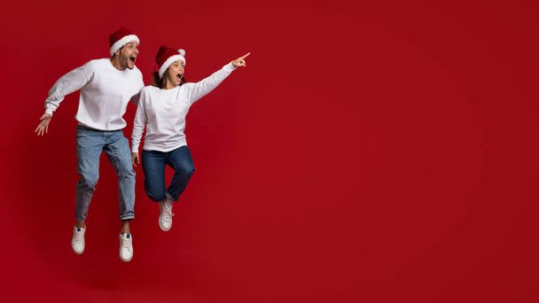 Удивительное предложение. Удивлённая молодая пара в шляпах Санта-Клауса, прыгающая и указывающая в сторону - Фото, изображение