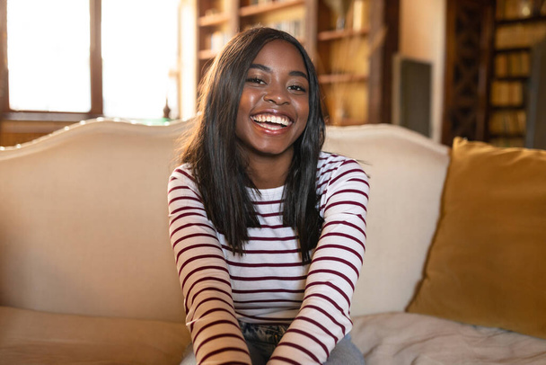 Porträt einer glücklichen jungen Afroamerikanerin, die in die Kamera lächelt und zu Hause auf dem Sofa sitzt - Foto, Bild