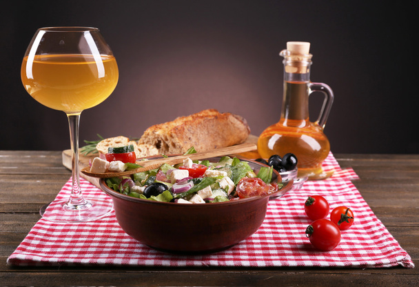 Bowl of Greek salad served with olive oil and glass of wine on napkin on wooden table on dark background - Photo, image