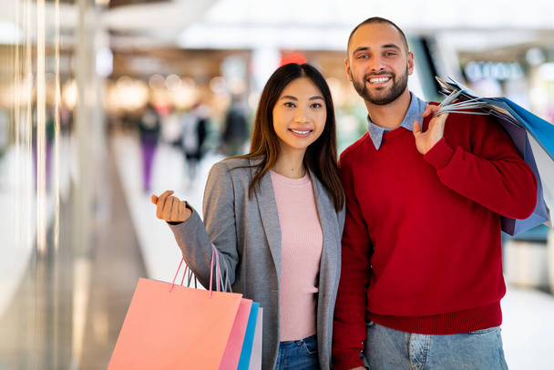 Feliz joven pareja multirracial con coloridas bolsas de comprador de pie juntos después de ir de compras en el centro comercial de la ciudad, espacio de copia - Foto, Imagen