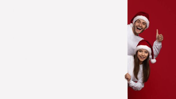 Ferienangebot. Fröhliches Paar in Nikolausmützen blickt hinter weiße Werbetafel - Foto, Bild
