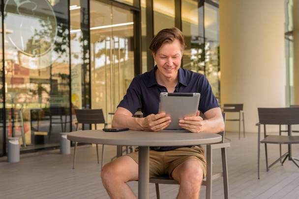 Hombre sentado al aire libre en la cafetería durante el verano mientras sonríe y el uso de la tableta digital de disparo horizontal - Foto, imagen