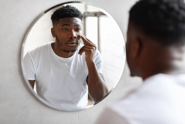Афроамериканец наносит крем-увлажнитель на лицо в ванной комнате - Фото, изображение