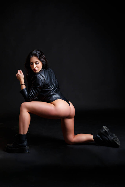 A slender tanned woman with a perfect body in a black bodysuit and a black leather biker jacket posing in the studio on a black background - 写真・画像
