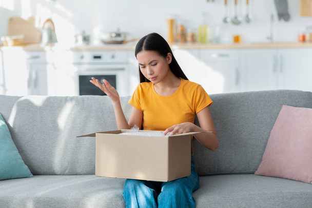 Wrong package. Upset asian lady sitting with box on sofa, sad about shipping mistake, displeased by bad delivery service - Fotoğraf, Görsel