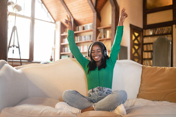 Carefree young black woman listening to music, using headphones, sitting on couch in living room, free space - Photo, image