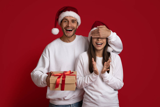 Nadšený mladý muž v santa klobouk zakrývající přítelkyně oči a dávat dárky - Fotografie, Obrázek