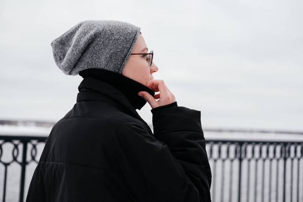 Mujer joven pensativa y pensativa con el pelo corto en sombrero, anteojos, abrigo negro caminando por una calle de la ciudad de invierno y mirando hacia otro lado. Disfrutando del clima y divirtiéndose. Gente cándida - Foto, imagen