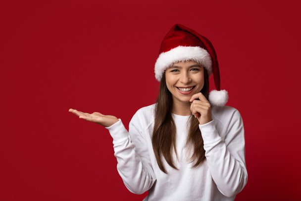 Xmas Offer. Cheerful Young Woman In Santa Hat Holding Invisible Object On Open Palm - 写真・画像