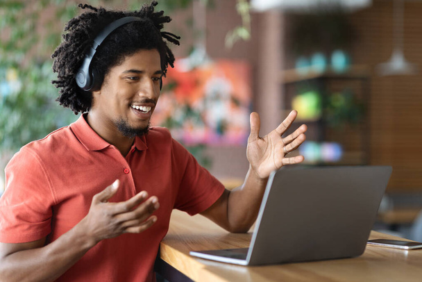 Νεαρός μαύρος άνδρας με ακουστικά που κάνει βιντεοκλήση με φορητό υπολογιστή στο καφέ - Φωτογραφία, εικόνα
