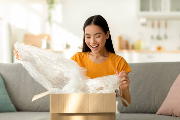 Fast delivery. Happy asian lady received package, unpacking cardboard box, sitting on the sofa at home, copy space - Photo, Image