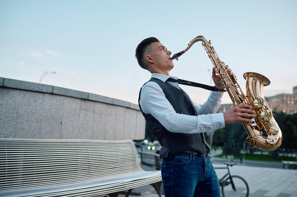 Saxophonist plays melody at the bench in city park - 写真・画像