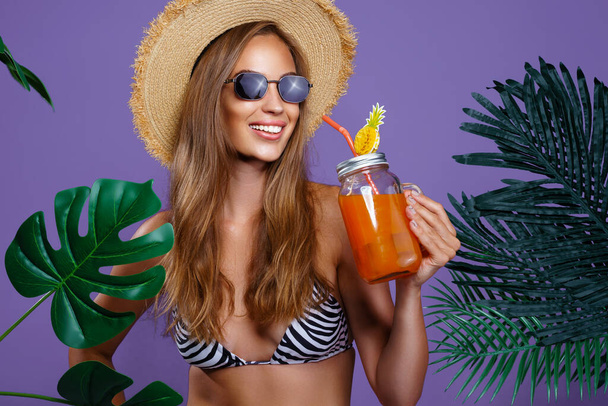 Happy beauty girl holds fruit cocktail in sunglasses and straw hat while stands near tropical plants - 写真・画像