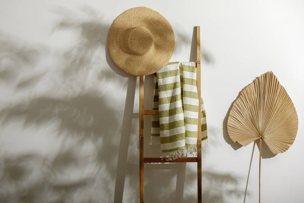 Organiczny ręcznik plażowy na drabinie drewnianej, słomkowy kapelusz i wysuszony liść palmowy na białej ścianie z cieniami z liści drzew - Zdjęcie, obraz