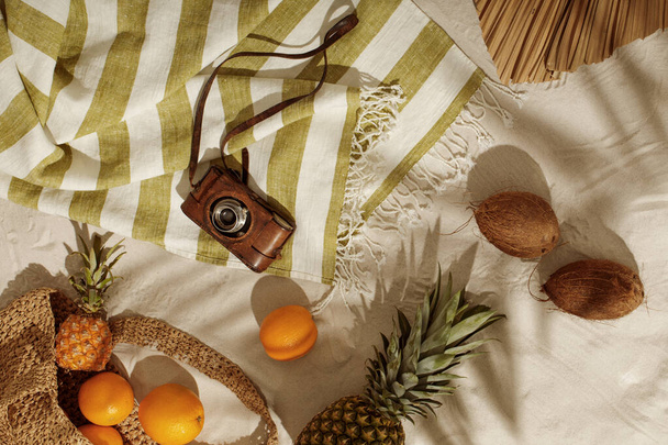 Fresh oranges, pineapple, coconuts falling out from woven bag on sandy beach. Vintage camera on linen beach towel with fringes. Tropical holidays concept - Foto, Imagem