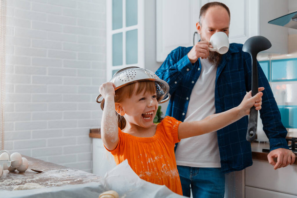 Happy daughter and father playing in kitchen in morning. Child kid girl put colander bowl on her head with ladle in hand she have fun and laughs Plays game. Happy time together family in cooking time - Photo, image