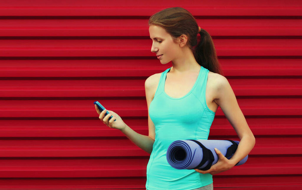 Fitness young woman with smartphone and yoga mat on red background - Photo, Image