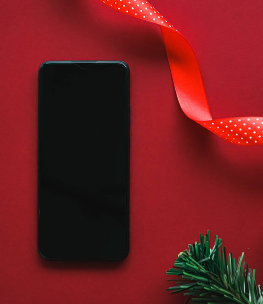 Χριστουγεννιάτικη εφαρμογή τηλεφώνου και μήνυμα διακοπών έννοια. Smartphone με λευκή μαύρη οθόνη και διακόσμηση Χριστούγεννα σε κόκκινο φόντο ως επίπεδη lay mockup σχεδιασμό - Φωτογραφία, εικόνα
