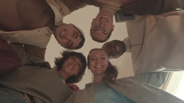 Widok z dołu ujęcie pięciu wesołych wielonarodowych studentów uniwersytetów przytulających się i uśmiechających stojąc razem w kręgu - Materiał filmowy, wideo