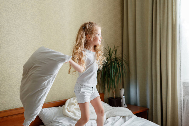 Cheery little Kid girl having fun in bedroom playing with pillow. Preschool daughter Caucasian blonde girl play in pillows cushions fight in bedroom on bed in home interior. - Photo, image
