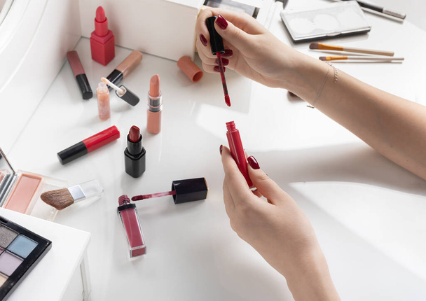 open red lip gloss in hands of girl on white dressing table against background of other lipsticks. Lipstick and lip gloss concept for makeup - Foto, afbeelding