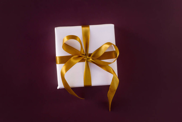 Caixa de presente embrulhada em papel branco com um arco dourado em fundo violeta festivo - Foto, Imagem