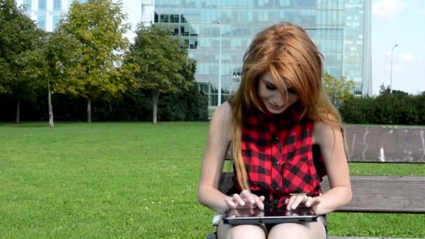 Mujer atractiva joven trabaja en la tableta (chat) y sonríe - se sienta en el parque (naturaleza - hierba y árboles) - edificio moderno
 - Imágenes, Vídeo