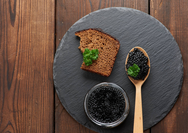Musta melokalakaviaari puulusikassa, leipäviipale ja purkki mustalla pyöreällä grafiittilevyllä, ylhäältä katsottuna - Valokuva, kuva