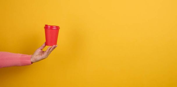 hand holds paper cardboard red cup for coffee, yellow background. Eco-friendly tableware, copy space - Photo, Image
