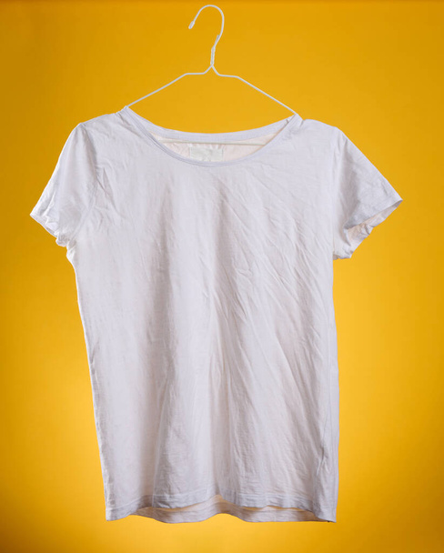 white cotton crumpled t-shirt hanging on a hanger on a yellow background, summer clothes - Photo, Image