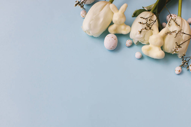 Buona Pasqua! Eleganti uova di Pasqua, tulipani e coniglietti piatti giacevano su sfondo blu. Moderne uova di Pasqua al cioccolato e disposizione dei fiori bianchi. Modello di biglietto di auguri, spazio per il testo - Foto, immagini