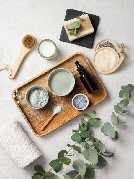the concept of organic cosmetics with clay, sea salt, soap,  brush and eucalyptus branch in a wooden tray on a white textured background. Top view. - 写真・画像